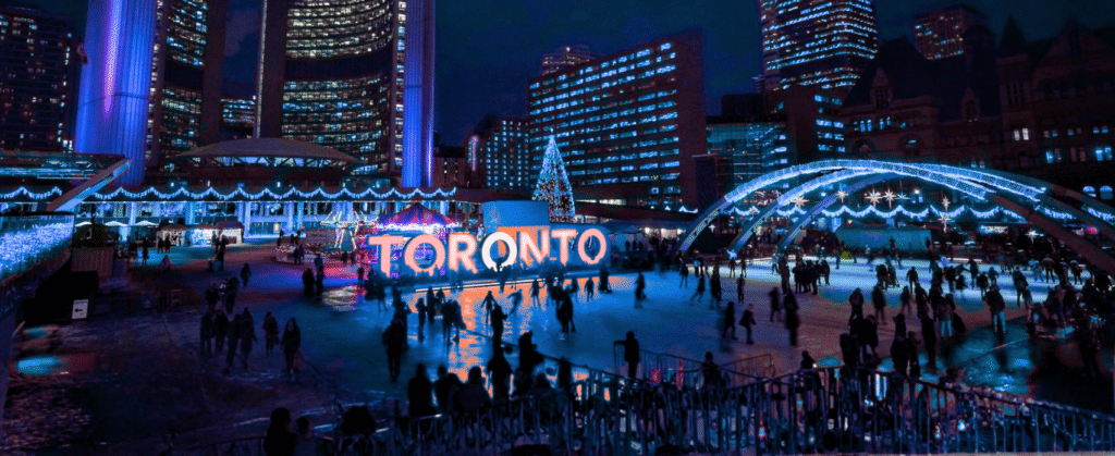 “Downtown Toronto at night featuring Nathan Phillips Square, TORONTO sign, and skating rink during a festive winter event.”
