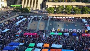 Aerial view of a large multicultural festival in Toronto with thousands of attendees, vendor tents surrounding the reflecting pool at Nathan Phillips Square, and the Toronto sign visible during a summer community event.
