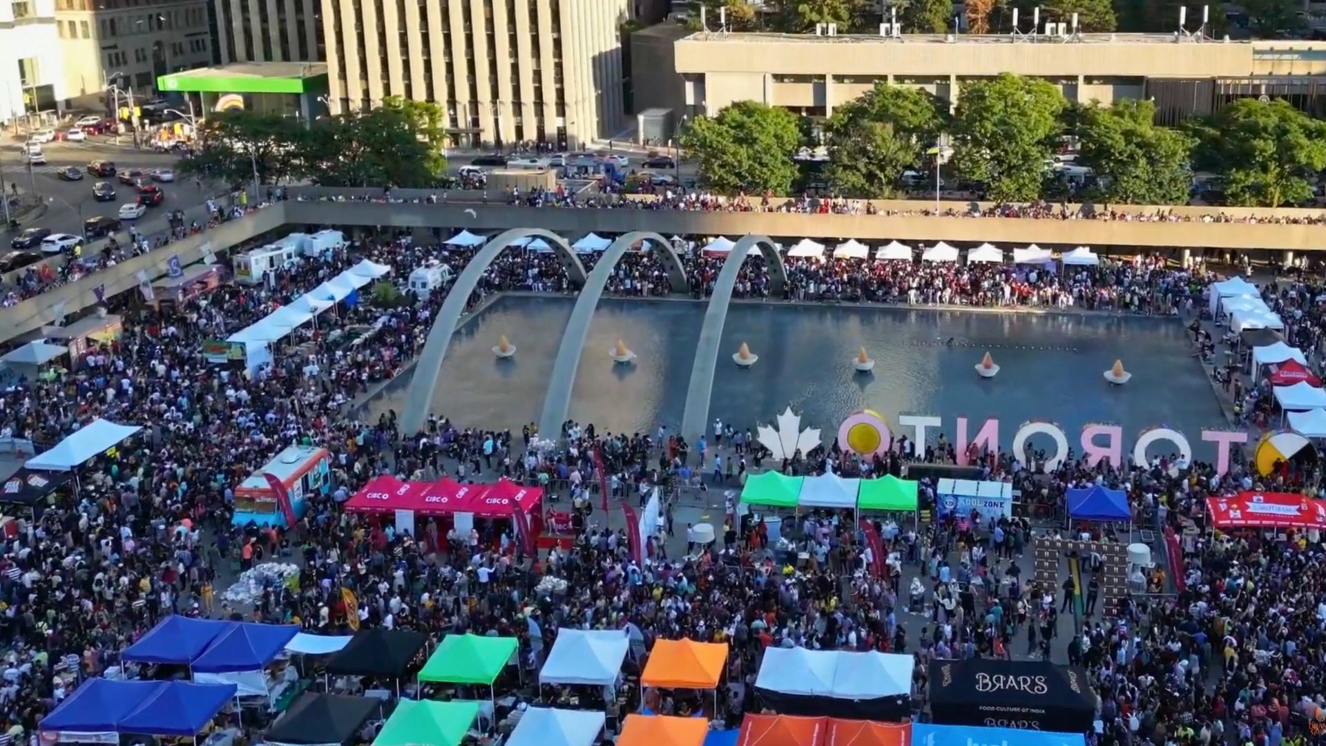 Aerial view of a large multicultural festival in Toronto with thousands of attendees, vendor tents surrounding the reflecting pool at Nathan Phillips Square, and the Toronto sign visible during a summer community event.