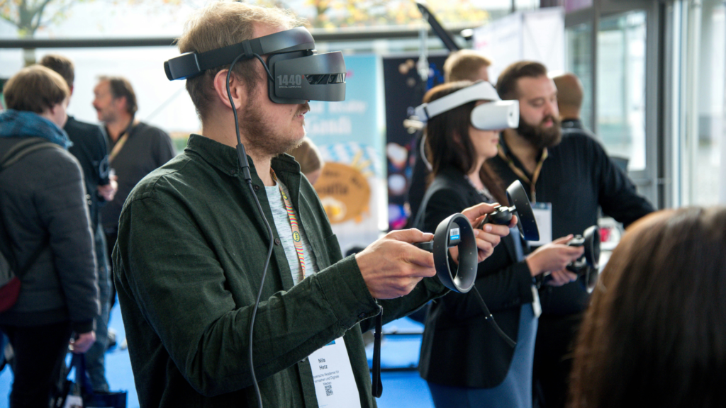 People at a tech event using virtual reality headsets and handheld controllers, engaging with immersive experiences while others observe in the background.