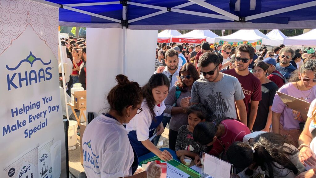 Crowded outdoor event booth where brand ambassadors offer samples to attendees gathered under a canopy.