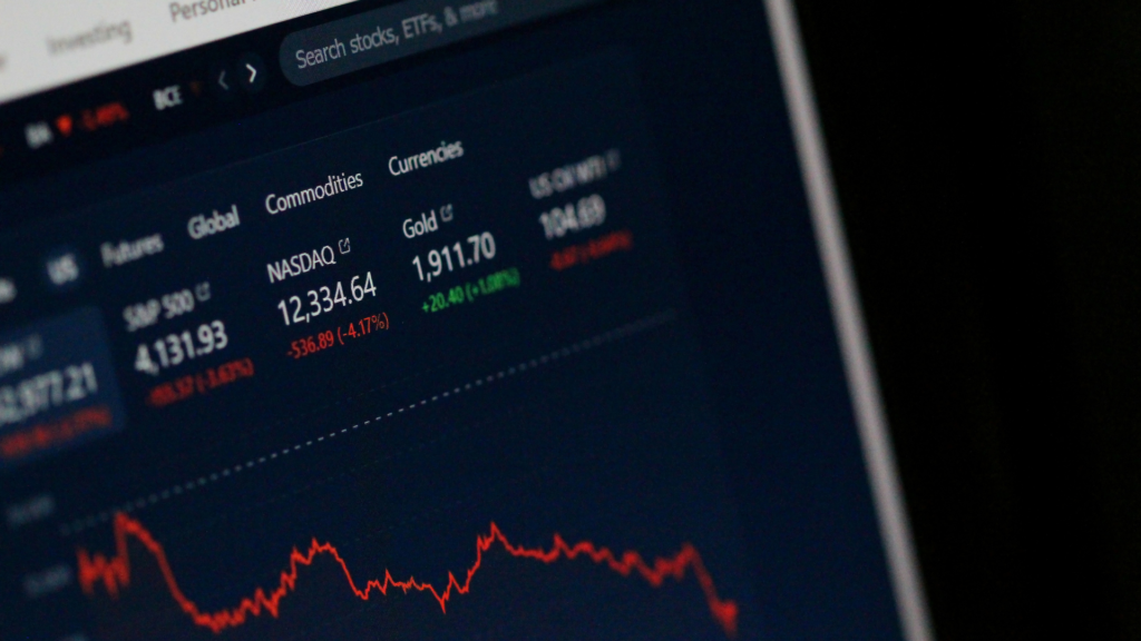 Close-up of a digital financial dashboard displaying stock market data, including NASDAQ, gold prices, and a downward-trending chart on a dark interface.