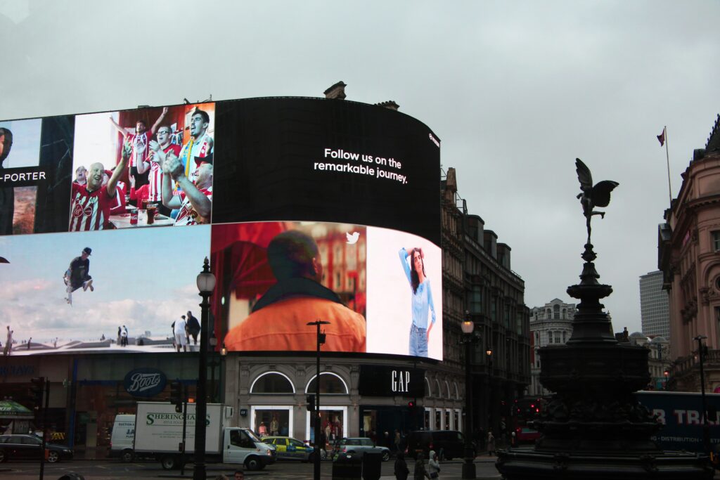 Large curved digital billboard in a busy city square displaying multiple brand visuals and a message reading “Follow us on the remarkable journey,” with surrounding urban architecture and street activity.