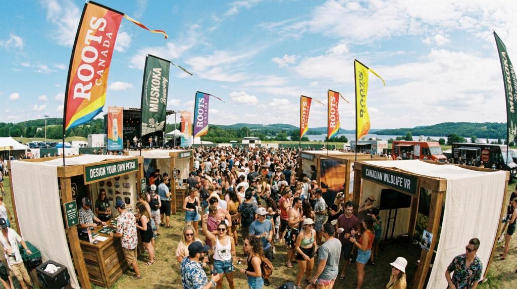 Busy outdoor brand activation festival with Roots Canada booths, interactive experiences, live music stage, and large crowd near waterfront.