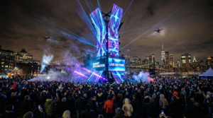 Nighttime experiential brand event with massive LED structure, drone light show, lasers, and large crowd by city skyline, showcasing immersive live activation experience.