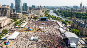 Aerial view of large outdoor festival in downtown Ottawa with main stage, thousands of attendees, vendor tents, and Ottawa River with Parliament buildings in the background.