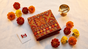 Punjabi community marketing Canada 2026: an overhead editorial flat-lay of Punjabi Canadian cultural identity objects — a phulkari-embroidered textile swatch in vibrant red, orange, and gold, scattered marigold flower heads, a small brass diya, and a mini Canadian flag pin.