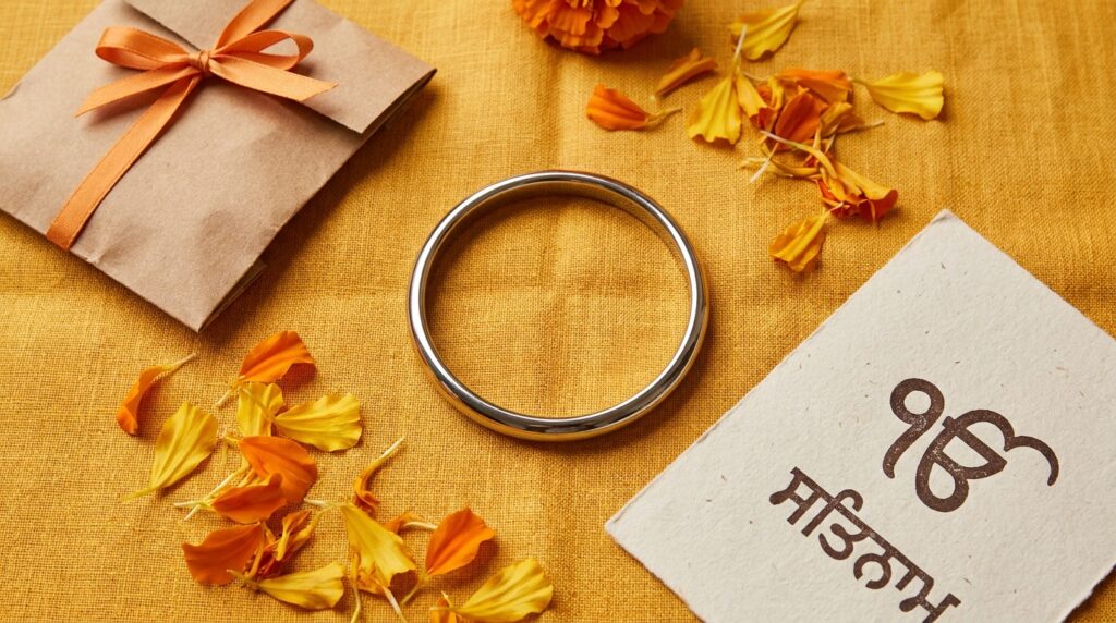 Punjabi community marketing Canada 2026: a close-up editorial flat-lay of Vaisakhi brand activation objects — a steel Sikh kada bangle, scattered marigold petals, an unbranded product sample package in vibrant orange, and a printed Gurmukhi script card.